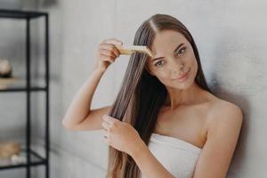 Young brunette woman brushes hair with comb after taking shower and applying hair care mask, wears minimal makeup, has healthy glowing skin after hygienic procedures, stands wrapped in towel photo