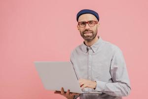 Photo of guy internet user browses website on laptop computer, connected to wireless internet, holds electronic gadget, wears spectacles, formal shirt, isolated over pink background with free space
