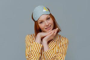 Portrait of good looking woman gets enough amount of sleep, enjoys pleasant relaxation, tilts head and keeps hands under chin, wears blindfold and striped pajamas, isolated over grey background photo