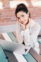 Indoor portrait of young successful Caucasian woman blogger writing a new post for her popular blog using notebook while sitting at desk over brick wall background. Smiling pretty lady with red nails photo