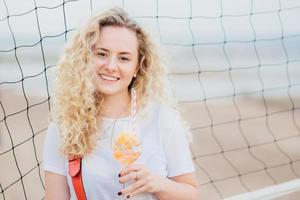 Adorable glad young European woman has bushy curly hair, smiles gently, holds glass of orange cocktail, stands near tennis net with copy space right for your advertisement or promotional text photo