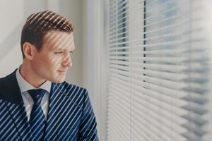 Indoor shot of pensive male economist dressed in formal outfit, looks pensively at window with jalousie conemplates about work during dinner break feels tired after working. Business, people, thoughts photo