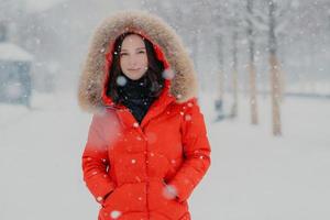 Lovely female model in red jacket, stads outdoor during snowfall, looks with dark eyes at camera, going to have stroll with boyfriend, breathes fresh air. People, winter time and free time concept photo