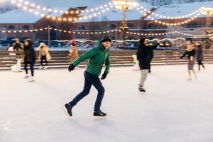 Outdoor shot of attractive man with beard, wears warm winter clothes, practices going skating on ice skate ring decorated with lights, has glad expression, enjoys his favourite action photo