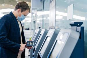 Sideways shot of man poses at self check in desk in airport, holds passport, going to register for flight, wears medical mask for safe traveling during coronavirus outbreak. Pandemic in public place photo