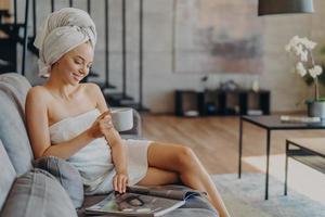 Horizontal shot of relaxed satisfied woman has healthy skin reads magazine poses with cup of coffee or tea at couch wrapped in soft towels enjoys spending time at home. Refreshment leisure spa photo