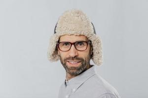 Close up shot of attractive male with stubble, wears glasses and fur hat, prepares for winter, has gentle smile models against white background. Masculine guy has blue eyes thick bristle stands indoor photo