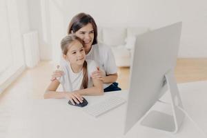 Modern technology and family concept. Caring lovely mother teaches little daughter how to use internet, explains how to download application, pose in spacious room with white walls, spend time at home photo