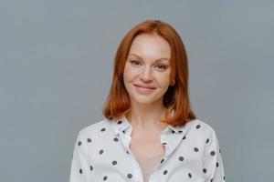 Portrait of attractive ginger European woman smiles gently at camera, wears makeup, dressed in polka dot blouse, looks directly at camera, models against grey background, thinks about changing job photo