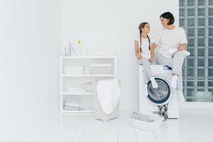 Housewife and small child sit on top of washing machine, holds freshly washed towels, do washing together, wear white t shirts, pants and socks, white console with iron, folded linen and detergents photo