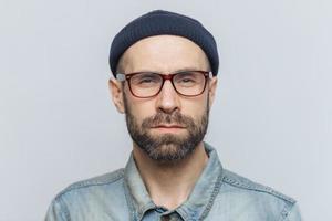 Photo of intelligent confident stylish man with dark thick beard and mustache, looks seriously into camera, poses against grey studio background, wears spectacles and hat. Facial expressions concept