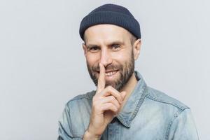 Cheerful attractive male with blue eyes shows silence sign, has satisfied expression, wears fashionable hat and denim jacket, isolated over grey background. Handsome man shows hush sign. Be quiet. photo