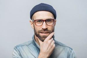 Confident satisfied bearded male keeps hand on chin, listens attentively interlocutor, wears fashionable clothing, isolated over white studio background. People and facial expressions concept photo