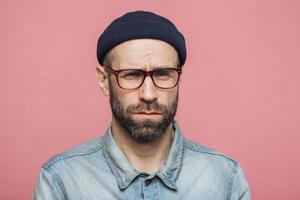 Unhappy discontent unshaven male looks with grumpy expression into camera, wears glasses black hat and stylish denim shirt, isolated over pink background. Displeased middle aged man poses indoor photo