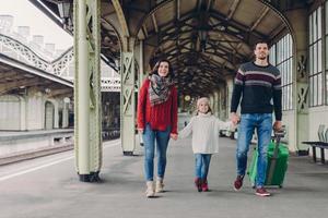 una foto de una familia feliz que va a tener un buen viaje durante las vacaciones, llevar una bolsa, caminar en la plataforma de la estación de tren, estar de buen humor. padre, madre e hijo llegan de viaje. concepto de viaje