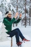 el retrato vertical de un hombre de aspecto agradable hace fotos con un teléfono inteligente, toma fotos de hermosos paisajes invernales, disfruta de la naturaleza y el clima helado, tiene una sonrisa agradable. personas, concepto de tecnología