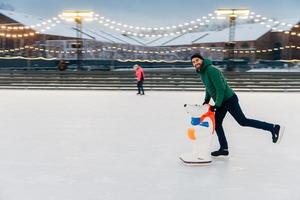 Portrait of happy middle aged male skater stands on ice ring, enjoys free time, has active lifestyle, being on ice arena. Smiling joyful man rejoices frosty winter weather. Skate aid. Hobby concept photo