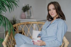Sideways shot of pretty woman sits crossed legs in wicker chair, dressed in pyjamas, drinks hot coffee and latte, poses against cozy interior indoor, has weekend. People and relaxation concept photo