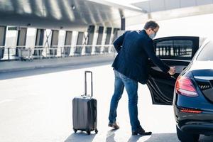 Man arrives from abroad, wears medical mask, returns from country with coronavirus outbreak, needs quarantine and self isolation, opens doos of car, places luggage in transport. Virus spread concept photo