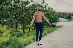 Sporty man with thick beard does exercises with jumping rope, has muscular torso, stays in good physical shape, poses outdoor. Fitness and healthy concept. Male runner warms up with sport equipment photo