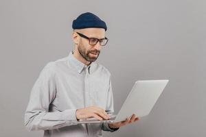 Professional IT developer downloads files, chats online in social networks, bloggs and surfes internet webpages wears formal shirt, stands against grey background. Technology and people concept photo