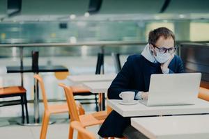 Man freelancer wears face mask to prevent spread of coronavirus and coughing, uses laptop computer for distant work, poses in cafe with cup of coffee, reads information about symptoms of disease photo