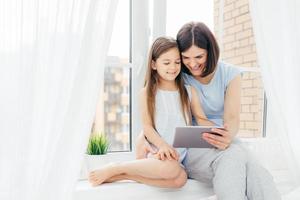 People, technology, family, children concept. Positive young other and her small daughter sit on window sill, hold digital tablet, watch interesting cartoon, pose near window with white curtains photo