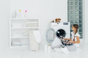 Housekeeping, children and domestic chores concept. Happy kid unloads washing machine, puts clean washed clothes in basin, curious dog looks from above, lies on washer in laundry room at home photo