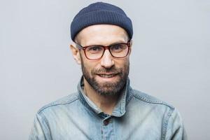 Indoor shot of handsome unshaven male with satisfied expression, rejoices achieving success at work, looks happily through spectacles, isolated over white background. Smiling bearded man indoor photo