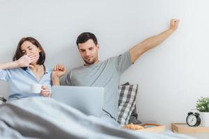 Family spend time together in bedroom, stretch and yawn as get up early, do remote work on laptop computer, use free wifi connection, drink coffee, being busy. Morning and awakening photo