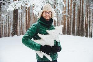 atractivo hombre barbudo con apariencia atractiva, usa anorak verde y sombrero, sostiene un abeto artificial, pasa los fines de semana en un clima helado en el bosque de invierno. concepto de invierno, clima y estilo de vida foto