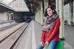 Glad young European woman wears red sweater, scarf, holds mobile phone, sits at bag, enjoy travelling, poses at railway station, smiles happily. People, tourism, travelling and technology concept photo
