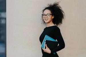 Sideways shot of beautiful dark skinned curly college student dressed in black casual outfit, carries textbook, notepad, smiles positively, isolated on grey background. People and studying concept