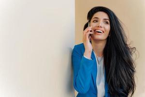 joven mujer de negocios positiva sonríe alegremente, se comunica por teléfono móvil, se viste formalmente, se enfoca en algún lugar, tiene un maquillaje mínimo. empresaria alegre resuelve problemas de trabajo de buen humor foto