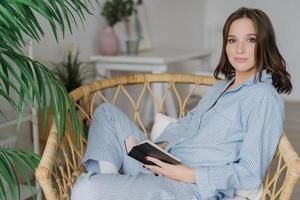 Sideways shot of young female reader hold book in hands, rests at home, wears pyjamas, enjoys calm atmosphere, reading bestseller, sits in wicker chair. People, hobby and relaxation concept. photo