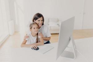 Indoor shot of happy lovely mother and daughter watch funny cartoon on computer, have pleasant smiles, enjoy togetherness, spend free time or weekend at home, spacious light empty living room photo