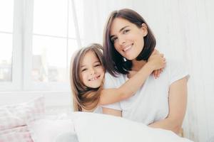 Positive daughter and mum embrace each other, being in high spirit after good sleep, pose in bedroom, smile gently on face, have pleasant appearance. Pretty child with her mother. Morning and bed time photo