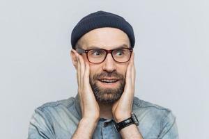 Amazed handsome middle aged man looks into distance in bewilderment, keeps hands on cheeks, wears glasses and hat, isolated over white background. People, emotions and unexpectedness concept photo