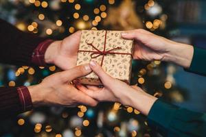 Wrapped box in womans and mans hands. Affectionate husband congratulates his wife with New Year, presents her holiday gift box, stand against decorated Christmas tree. Holiday concept photo