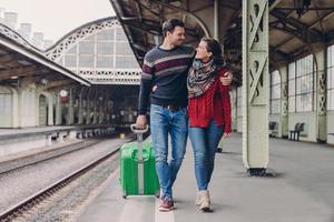 Young family couple embrace and look at each other with smile, wear jeans and warm sweaters, carry suitcase, pose on railways station meet after long departure. Boyfriend and girlfriend cuddle outside photo