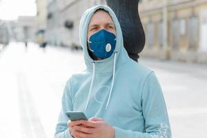 Horizontal outdoor shot of young man wears respirator mask for health protection, walks on street with mobile phone, protects himself during coronavirus outbreak, follow news online, dressed in hoodie photo