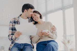 toma interior de una mujer relajada y hermosa con una taza de café, recibe un abrazo de su esposo que tiene papeles, hacen un informe financiero juntos, tienen su propia empresa comercial. concepto de familia, finanzas y presupuesto. foto