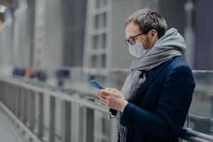 una foto lateral de un bloguero masculino envía mensajes de texto y lee comentarios de seguidores en teléfonos celulares modernos, usa una máscara médica estéril para protegerse del coronavirus u otras enfermedades infecciosas