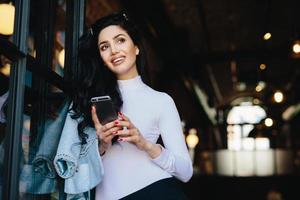 Dreamy stylish brunette lady with appealing appearance dressed elegantly holding mobile phone in hands watching photos remembering pleasant moments in her life. Woman with pensive expression