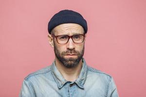 Grumpy bearded man with offended expression, being dissatisfied with something, frowns face, wears spectacles, hat and shirt, isolated over pink background. Displeasure and negativity concept photo