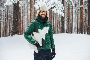 Cheerful bearded male wears warm anorak and hat, walks in winter forest, holds artificial fir tree, has festive mood, going to celebrate New Year. Outdoor shot of male spends free time on cold weather photo