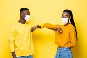 Young African American couple wearing face masks and bumping their elbows for greeting during COVID-19 pandemic in isolated yellow studio background photo