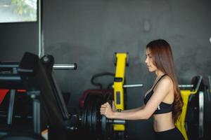 Fit young woman lifting barbells looking focused, working out in a gym with other people photo