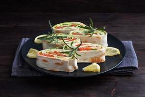 Rolls of thin pita bread and red salted salmon with lettuce leaves on a black ceramic plate, dark wooden background. Copy space. photo