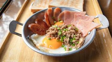 huevos fritos en una sartén pequeña con jamón, cerdo picado, cebolleta y salchichas chinas en bandeja de madera. desayuno tailandés popular llamado kai kata foto